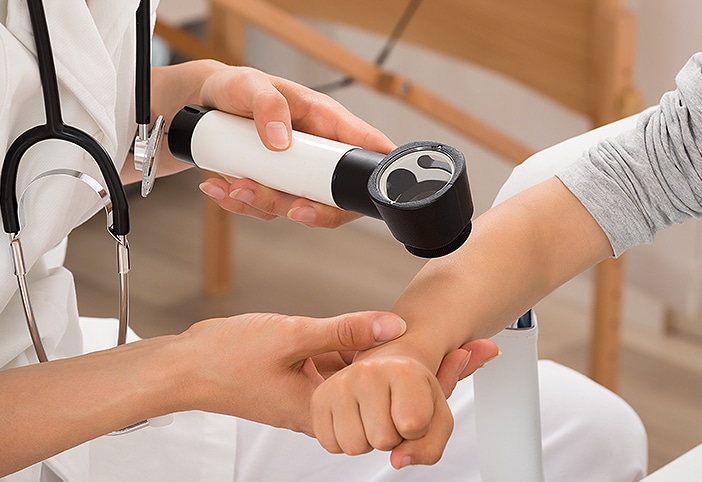 Stock photograph of models: a masked clinician in a hair cover checks a reclining woman’s forehead with a dermatoscope